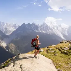 A hiker with a red backpack pauses on a rocky outcrop, enjoying the breathtaking view of the Mont Blanc massif during a challenging hike.