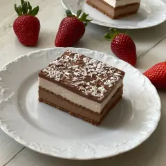 Pyszne ciasto na zimno z warstwami czekolady i śmietanki, posypane wiórkami kokosowymi. Obok leżą świeże truskawki.