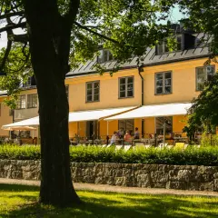 En gul byggnad med vita markiser och uteservering. En lång rad med träd skymmer delvis vyn.