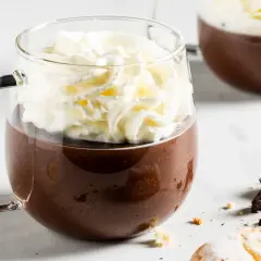 Una tazza di fumante cioccolata calda coronata da panna montata, con pezzi di cioccolato e biscotti sullo sfondo.