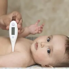 Dłoń rodzica mierzy temperaturę niemowlaka. Maluch leży spokojnie, patrząc w obiektyw.