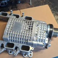 Srebrna sprężarka mechaniczna z kolektorem dolotowym, część do Mercedes Benz C32 AMG, leży na kartonie.
