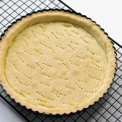 Base di pasta sucrée dorata e forata, pronta per essere farcita, su una griglia nera.