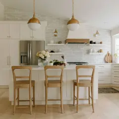 A bright, airy modern farmhouse kitchen with white subway tile, gold pendant lights, and a large island with three wooden stools.