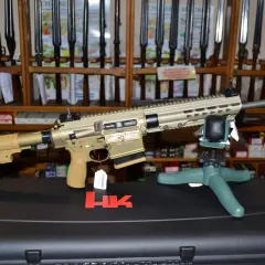 Un fusil HK MR 308, de couleur sable, est présenté sur un support. Derrière, des étagères chargées de fusils et de boîtes de munitions.
