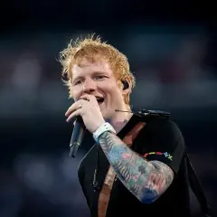 Ed Sheeran śpiewa z mikrofonem, jego ręka ozdobiona kolorowymi tatuażami.
