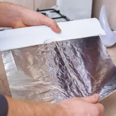 Ręce trzymają biały dozownik, z którego wysuwa się matowa strona folii aluminiowej. Gotowanie w kuchni.