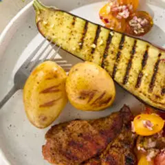 Une demi aubergine au barbecue, des pommes de terre grillées et une salade de tomates sur une assiette blanche.