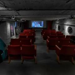 Kameralna sala kinowa z czerwonymi fotelami i zabytkowym projektorem. W tle ekran z filmem i dekoracje nawiązujące do historii kina, jakby to było pierwsze kino w Polsce.