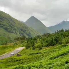 A car drives on a winding road through lush green Scottish Highlands, perfect for a hiking itinerary.