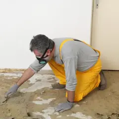 Un homme en salopette jaune et masque retire la colle de moquette avec une spatule. Une méthode naturelle pour enlever colle moquette.