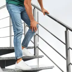 Mann mit Jeans und grünem Shirt hält sich das Knie, während er eine Treppe hinuntergeht. Er hat Probleme beim Treppen runter gehen.