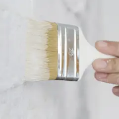 Pennello bianco stende pittura su muro grezzo. Si può mischiare il fissativo nella pittura per una finitura migliore.