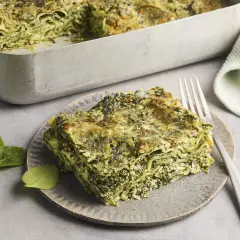 Eine Portion Spinat Ricotta Lasagne auf einem Teller, daneben eine Auflaufform mit der restlichen Lasagne. Frische Spinatblätter liegen daneben.