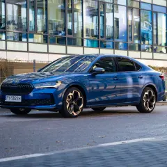 Elegancka, błękitna Skoda Superb 2025 z nowoczesnymi felgami stoi przed szklanym budynkiem.