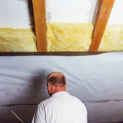 Mann dichtet ein altes Dach von innen ab, indem er Dämmmaterial zwischen den Holzbalken anbringt und eine weiße Folie spannt.