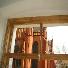 Widok przez stare okna skrzynkowe na ceglaną, zabytkową budowlę z wieżami i ozdobnymi detalami.