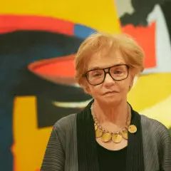 A woman with glasses and a statement necklace stands before a vibrant abstract expressionist painting.