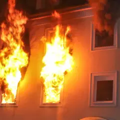 Flammen schlagen aus Fenstern eines Gebäudes. Dunkler Rauch steigt auf. Ein Beispiel für das richtige Verhalten im Brandfall ist die sofortige Evakuierung.