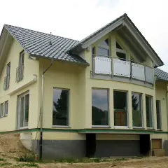 Nowy dom z żółtą elewacją i dachem w kształcie litery L. Duże okna i balkon dodają mu nowoczesnego charakteru.
