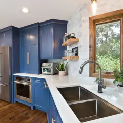 Stylish kitchen sink styling with a modern faucet, double basin sink, and a window overlooking greenery. Blue cabinets and stainless steel appliances complete the look.