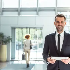 Uomo sorridente in abito scuro con tablet, in un moderno atrio di un pms hotel.