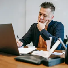 Zamyślony audytor siedzi przy biurku, pisząc coś na papierze i patrząc na ekran laptopa.