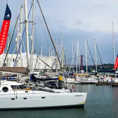 Una **barca lunga e sottile**, la Scarlett, è ormeggiata in un porto pieno di altre barche a vela. Sullo sfondo, un ponte rosso attraversa il fiume.