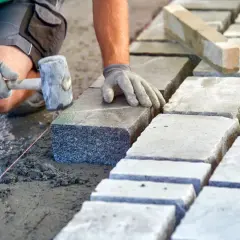 Układanie kostki brukowej na podsypce. Ręce w rękawiczkach kładą kamień, używając gumowego młotka. Widać suchy beton proporcje.