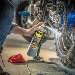 Persona en cuclillas engrasa cadena moto con spray WD-40.