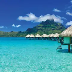 Bora-Bora loty: domki na wodzie w turkusowej lagunie, w tle zielona góra pod błękitnym niebem z chmurami.