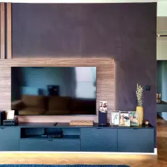 Nowoczesny salon z telewizorem, pod którym znajduje się szafka RTV. Ciepłe, drewniane panele i dyskretne oświetlenie LED nad telewizorem tworzą przytulną atmosferę.