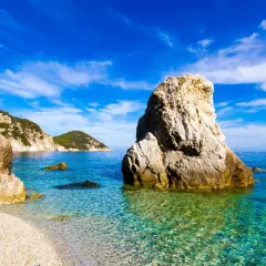 Plage de galets et rochers imposants dans une eau turquoise cristalline, évoquant une charmante ville toscane bord de mer sous un ciel bleu éclatant.