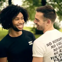 Due uomini sorridono, uno con maglia nera con simbolo di uguaglianza, l'altro con maglia bianca con messaggi di inclusione. Un'immagine che evoca il poliamore significato di amore libero e rispetto.