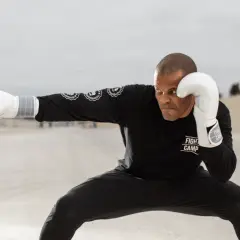 A boxer in a low crouching stance, wearing white gloves and a black "Fight Camp" shirt, throws a punch in an open, dusty landscape.