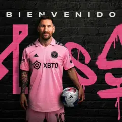 Lionel Messi w różowej koszulce Inter Miami, z piłką w ręku, na tle napisu "Bienvenido Messi". Nowy inter miami trener.