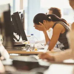 A woman sits at her desk, head in hands, looking demoralized at work. A bottle of water sits nearby.