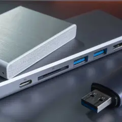 Ein silbernes Gehäuse für eine externe Festplatte liegt auf einem USB-Hub mit SD-Kartenleser und mehreren USB-Anschlüssen. Daneben ein USB-Stick.