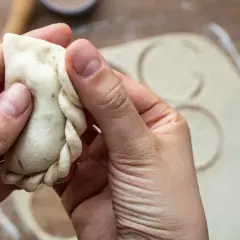Dłonie formują brzeg pieroga, pokazując, jak lepić pierogi. W tle rozwałkowane ciasto z wyciętymi kółkami.