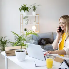 Uśmiechnięta kobieta prowadzi rozmowę telefoniczną, pracując zdalnie okazjonalnie przy laptopie. Na biurku napój i rośliny.