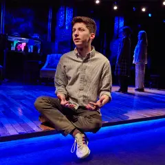 Andrew Barth Feldman sitzt im Schneidersitz auf einer Bühne, die in blaues Licht getaucht ist. Zwei Frauen stehen im Hintergrund.