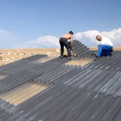 Zwei Männer verlegen graue Faserzementplatten auf einem Dach. Die Wellplatten werden auf einer Holzkonstruktion befestigt.