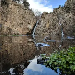 Cascadas gemelas caen entre rocas escarpadas en el embalse de Pedrezuela, reflejándose en el agua tranquila.