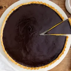 Ein Stück vom originalen Tarte au Chocolat Rezept wird mit einem goldenen Tortenheber herausgehoben. Daneben liegen Schokoladenstücke und eine Tasse Kakao.