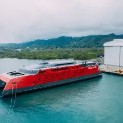 Nowy, czerwono-szary prom Fjord FSTR gotowy do rejsu do Norwegii. W tle zielone wzgórza i biała hala produkcyjna.