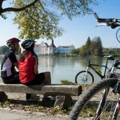 Para rowerzystów odpoczywa nad rzeką, podziwiając malownicze **trasy rowerowe Austrii** i pałac w oddali.