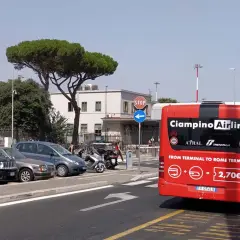 Czerwony autobus Ciampino Airlink czeka na pasażerów. Z tyłu widnieje napis "FROM TERMINAL TO ROME TERMINI" i cena 2,70€. W tle parking z samochodami i budynek lotniska.