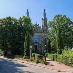 Neogotycki kościół w Krynkach, otoczony zielenią, z wieżami i charakterystycznym oknem.