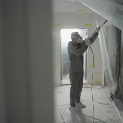 Pracownik w kombinezonie i masce ochronnej nakłada gładź agregatem. Pokój zabezpieczony folią.
