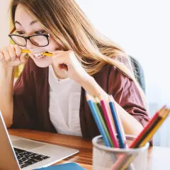 Dziewczyna z okularami gryzie ołówek, zastanawiając się, czy angielski jest trudny. Obok laptopa stoi koszyczek z kolorowymi kredkami.